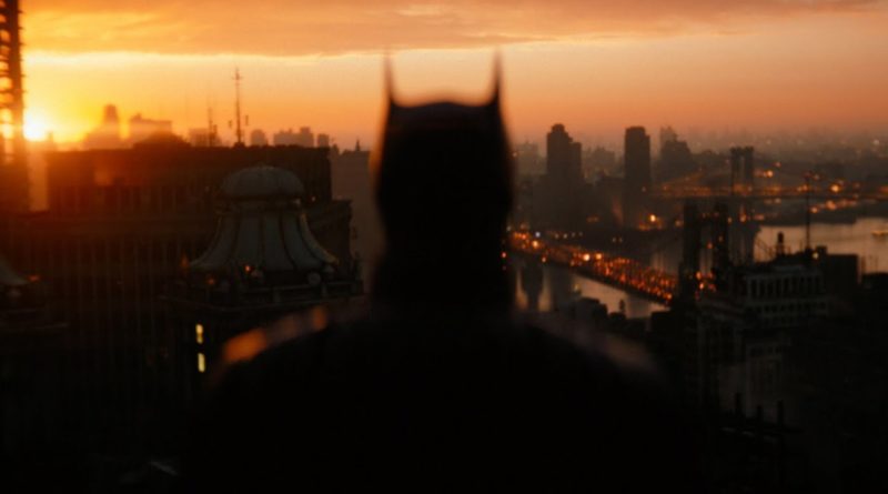 Batman is silhouetted against the coral and orange sky. His back is to the camera, and he is gazing down at Gotham from a rooftop.